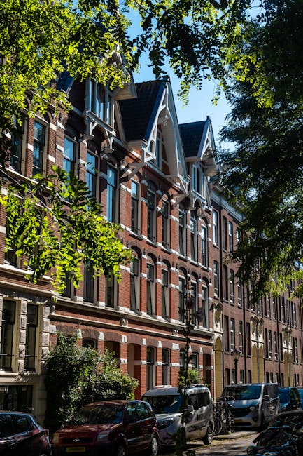 A row of multi-storey residential buildings with brick facades and white decorative trims, situated along a tree-lined street with parked cars visible at the curb. The buildings feature large windows, some with small balconies, and pitched roofs with gables. Sunlight filters through the leafy canopy overhead, casting shadows on the pavement below. The scene depicts an urban environment typical for home relocation or house removals, with a focus on the exterior architecture and street setting, suitable for the content on Holland Park Avenue moving guide for house removals by Holland Park Man and Van.