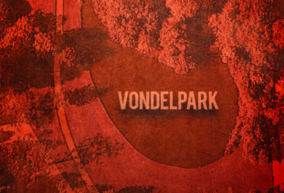 Aerial view of Vondelpark in Amsterdam, featuring a large oval-shaped lawn area surrounded by dense trees with reddish-orange foliage, possibly during autumn. A paved pathway runs alongside the park, with a few small pedestrians visible on the walkway. The park's name, 'VONDEL PARK,' is prominently displayed in large white capital letters within the grassy area. The scene is captured in warm, natural daylight, highlighting the contrast between the lush greenery and the vibrant tree colors. This image is relevant to house removals, as it depicts urban green spaces often encountered during residential relocations in the area, aligning with the context of furniture transport and home relocation logistics with [COMPANY_NAME] at hollandparkmanandvan.com, who handle removals within this environment.