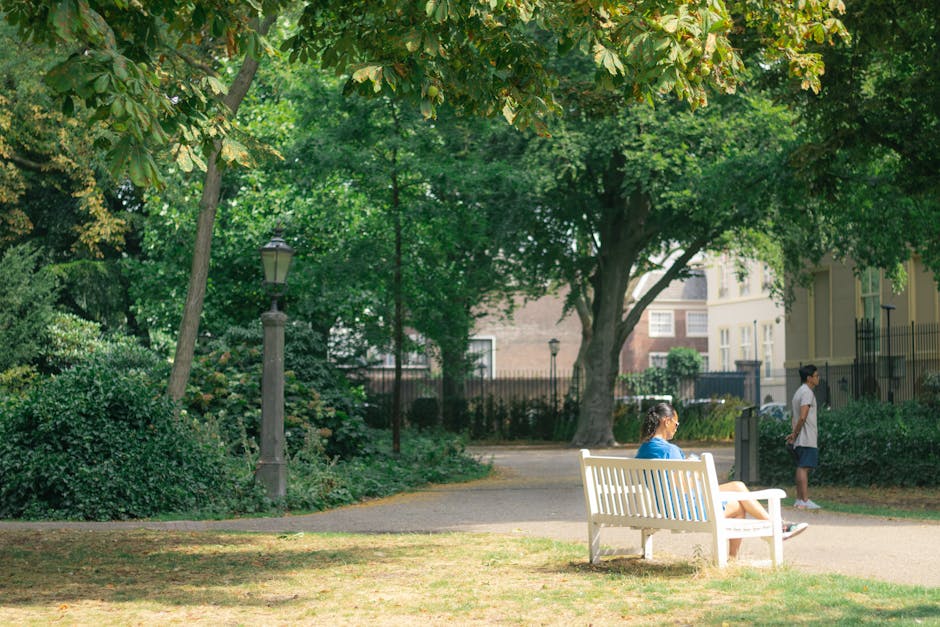 A woman is seated on a white park bench in an outdoor area with lush green trees and shrubs surrounding her. She is wearing a blue top and appears to be resting or waiting, with her legs crossed and her hands on her lap. Behind her, a young girl dressed in a white shirt and shorts stands near a black iron fence, looking towards the distance. The scene is set on a paved pathway within a residential area, with multi-storey buildings partially visible through the trees in the background. A vintage-style street lamp is situated along the pathway, and the area is illuminated by natural daylight, emphasizing the calm and organized environment typical for neighborhood leisure or casual outdoor activities. Holland Park Man and Van may use such scenic locations for local removals or home relocation services, highlighting their experience in moving within peaceful, residential neighborhoods.