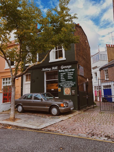 The image shows a two-storey building with a dark green facade and white lettering indicating it is the Notting Hill Garage, offering services such as servicing, repairs, diagnostics, and MOTs, located on a cobbled street with a tall tree partially obscuring the front. A classic grey car is parked directly in front of the garage entrance, which is open, revealing the interior workspace. To the right, a black metal gate leads into the property, and adjacent residential buildings with differing brick and painted exteriors are visible. The scene is illuminated by natural daylight, with a partly cloudy sky overhead. As part of house removal and furniture transport services, [COMPANY_NAME] may be involved in loading household items such as cardboard boxes, wooden furniture, and packing materials onto a van parked nearby, ready for a home relocation from Notting Hill Gate to Holland Park, ensuring careful handling and organized logistics in the loading process.