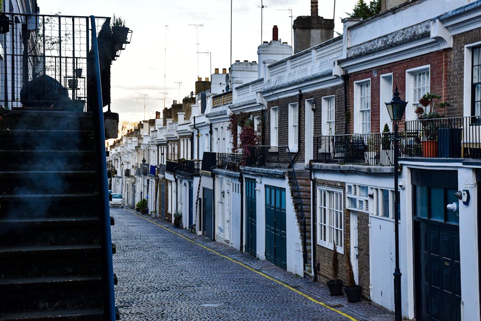 A row of terraced houses in a residential area with a cobblestone pavement in the foreground. The houses are multi-story with brick and painted exteriors, featuring small front gardens or balconies with potted plants and decorative railings. Some houses have external staircase access to upper floors, while others have front doors directly accessible from the pavement. A black lamppost is visible near one of the houses, and there are various windows, some with shutters. The image is taken during daylight with a partly cloudy sky, and the scene depicts an urban setting suitable for house relocations. The surroundings suggest a typical London street, often accessed for home relocation and furniture transport by services such as Holland Park Man and Van, which manage removals and moving logistics in the area.